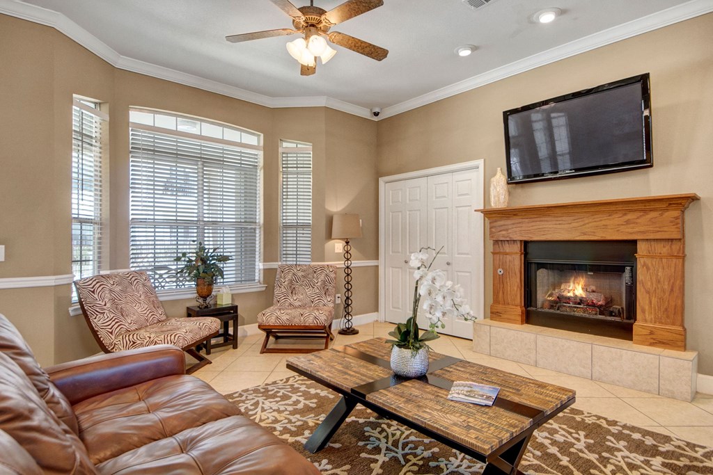 Cozy living room here at Mission Green Apartment Homes with a brown leather sofa, patterned armchairs, and a wooden coffee table. A fireplace, TV, and large window create a warm atmosphere.