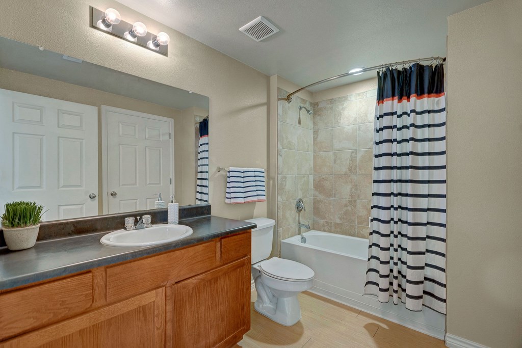 A modern bathroom here at Mission Green Apartment Homes features a wooden vanity with a dark countertop, a large mirror, and a striped shower curtain. Soft lighting creates a cozy ambiance.