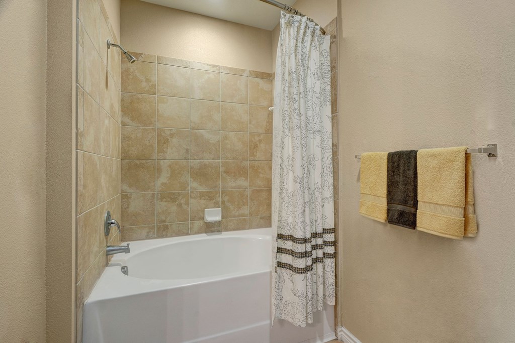 Bathroom here at Mission Green Apartment Homes with white tub, tan tiled walls, patterned shower curtain, and two folded towels on a rack. Neutral tones create a calm ambiance.