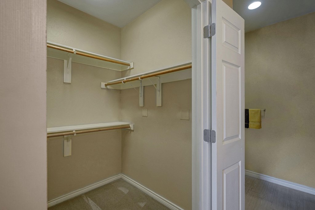 Empty beige walk-in closet here at Mission Green Apartment Homes with open door, wooden rods, and shelves. The room appears clean and organized, conveying a sense of spaciousness.