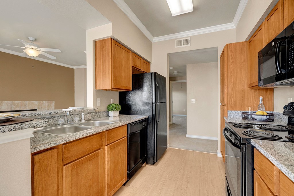 Modern kitchen here at Mission Green Apartment Homes with granite countertops, wooden cabinets, and appliances including a black stove, fridge, and microwave. Bright, clean, and inviting.
