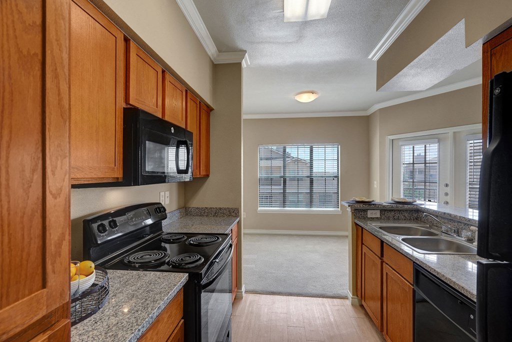 Bright kitchen here at Mission Green Apartment Homes with wooden cabinets, granite countertops, and black appliances, including a microwave and stove. A sunlit dining area with large windows is visible.