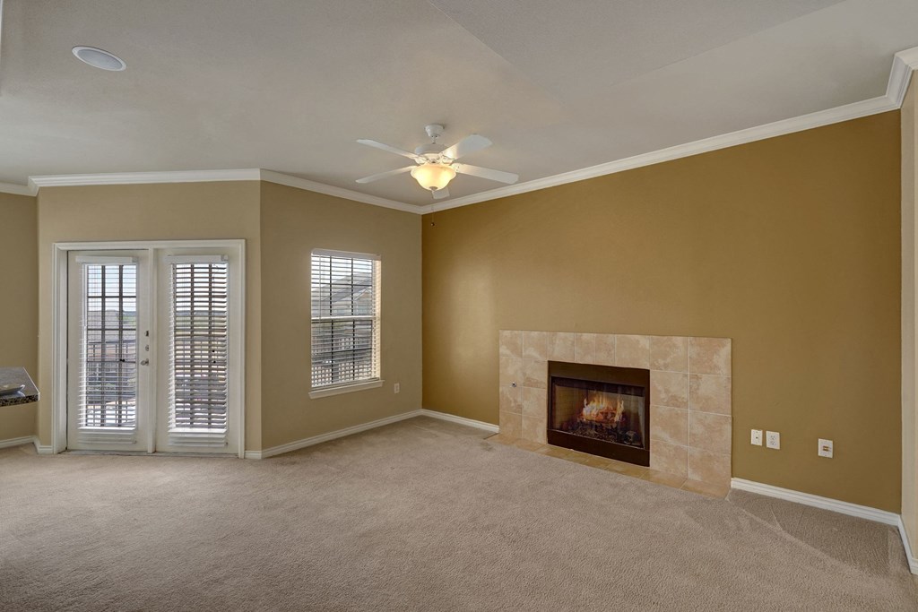 A cozy, vacant living room here at Mission Green Apartment Homes with beige carpet, tan walls, and a tiled fireplace. French doors and a window let in natural light. A ceiling fan hangs above.