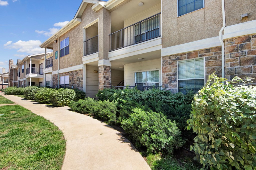Suburban Mission Green Apartment Homes with stone and stucco facade. A sidewalk curves through well-maintained shrubs under a clear blue sky, conveying a peaceful setting.