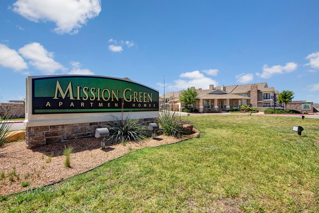 Sign reads "Mission Green Apartment Homes" against a bright, sunny backdrop. Nearby is a two-story building with landscaped grounds, conveying a welcoming atmosphere.