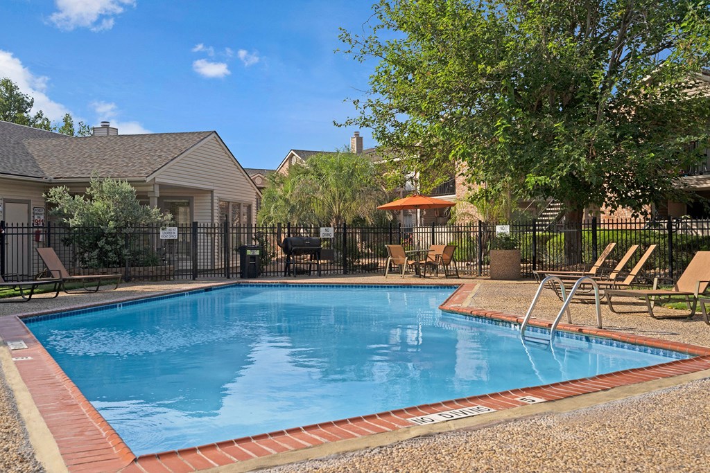 Outdoor pool here at Northridge Court Apartment Homes in a residential area with lounge chairs and trees surrounding it. A patio table with an orange umbrella is nearby, creating a relaxing atmosphere.