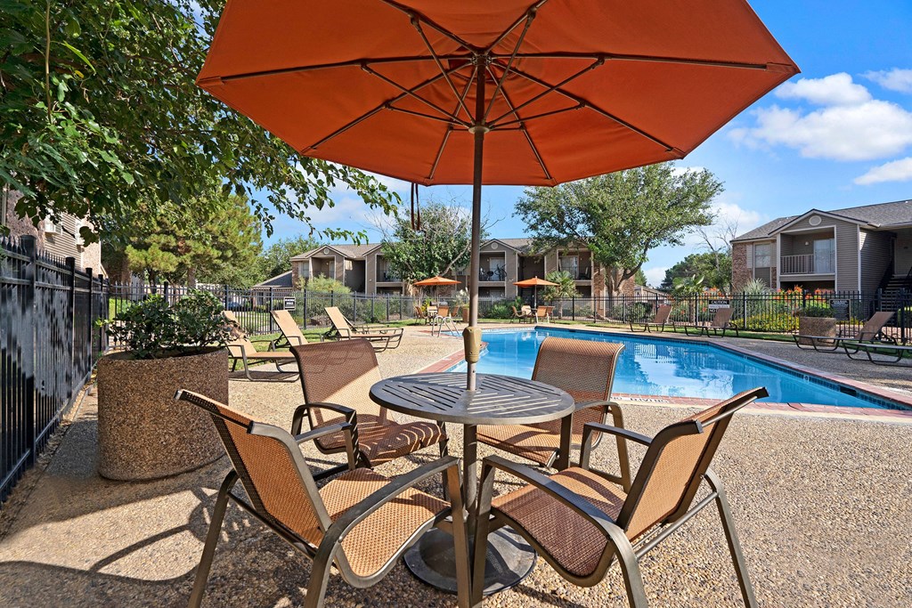 Outdoor pool area here at Northridge Court Apartment Homes with orange umbrellas and seating. Surrounded by apartment buildings and trees under a clear blue sky, conveying a serene atmosphere.