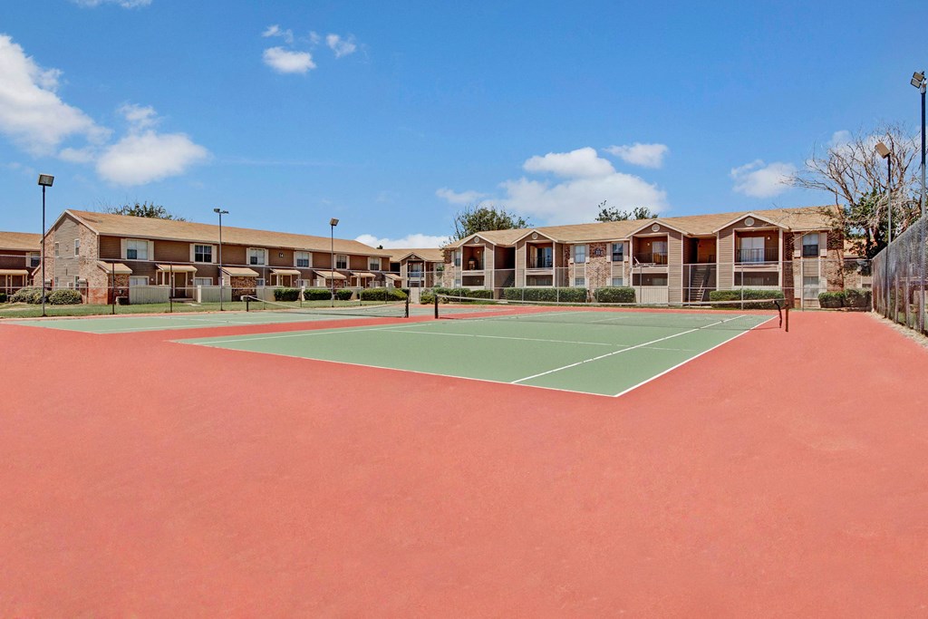 A tennis court here at Northridge Court Apartment Homes with a red surface and green playing area is surrounded by a chain-link fence. In the background, there are two-story brick apartments under a blue sky with scattered clouds.