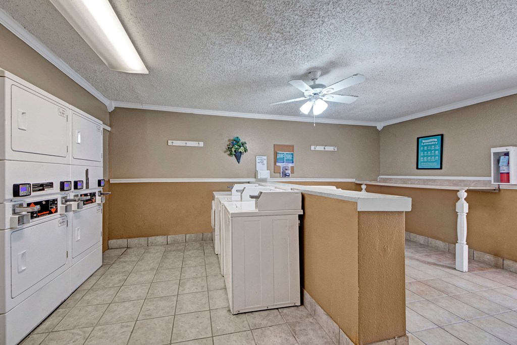 Laundry room here at Northridge Court Apartment Homes with beige walls and tiled floor, featuring white washing machines and dryers on the left. A ceiling fan is on, and wall decor includes a plant and framed sign, creating a clean, functional space.