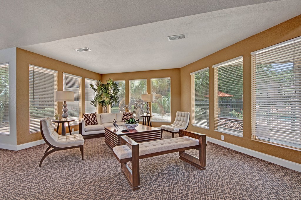 Bright, elegant lounge here at Northridge Court Apartment Homes with beige walls and large windows. Modern furniture includes white chairs and a wooden coffee table. Plants add a fresh touch.