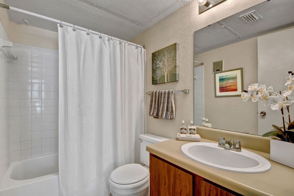 A bright bathroom here at Northridge Court Apartment Homes with a white shower curtain, wooden vanity, and beige countertop. Decor includes framed art, striped towel, and white orchid. Cozy and clean atmosphere.