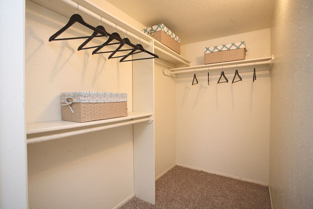 Empty walk-in closet here at Northridge Court Apartment Homes with beige walls and carpet. Black hangers on two racks and three decorative storage baskets on shelves convey tidiness and organization.