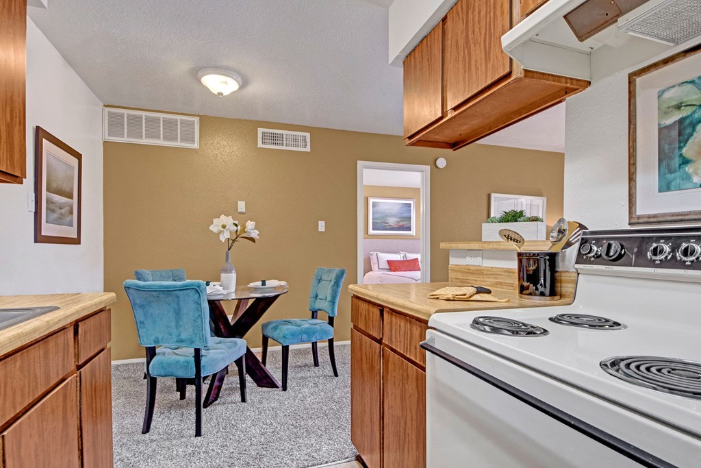 A cozy kitchen and dining area here at Northridge Court Apartment Homes with wooden cabinets, a white stove, and a small round table with blue chairs. Soft lighting adds a warm ambiance.