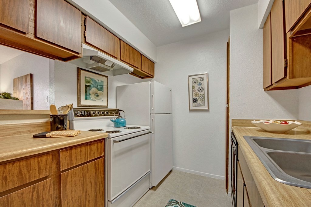 Compact kitchen here at Northridge Court Apartment Homes with wooden cabinets, white appliances, a stove with a turquoise kettle, and a dual sink. Warm, inviting atmosphere with artwork on walls.