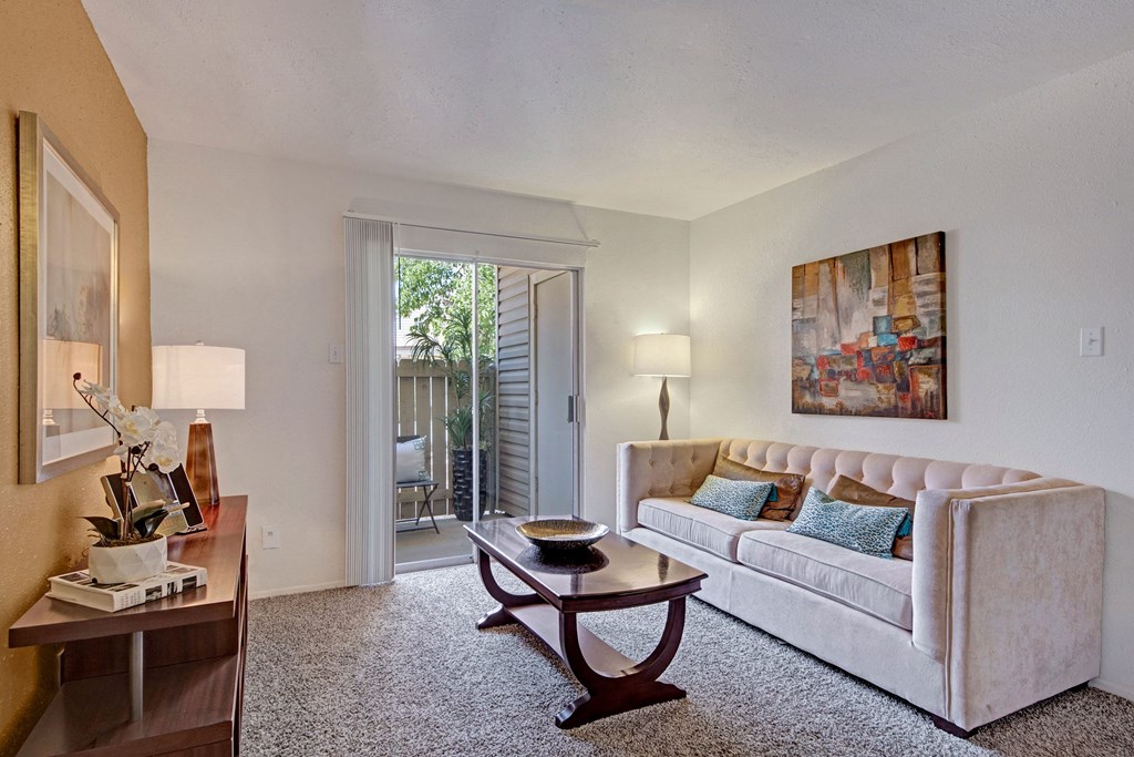 A cozy living room here at Northridge Court Apartment Homes with a beige sofa, blue cushions, and abstract art above. A wooden table stands on a carpet, with sliding doors opening to a patio. Soft lighting from lamps creates a warm ambiance.