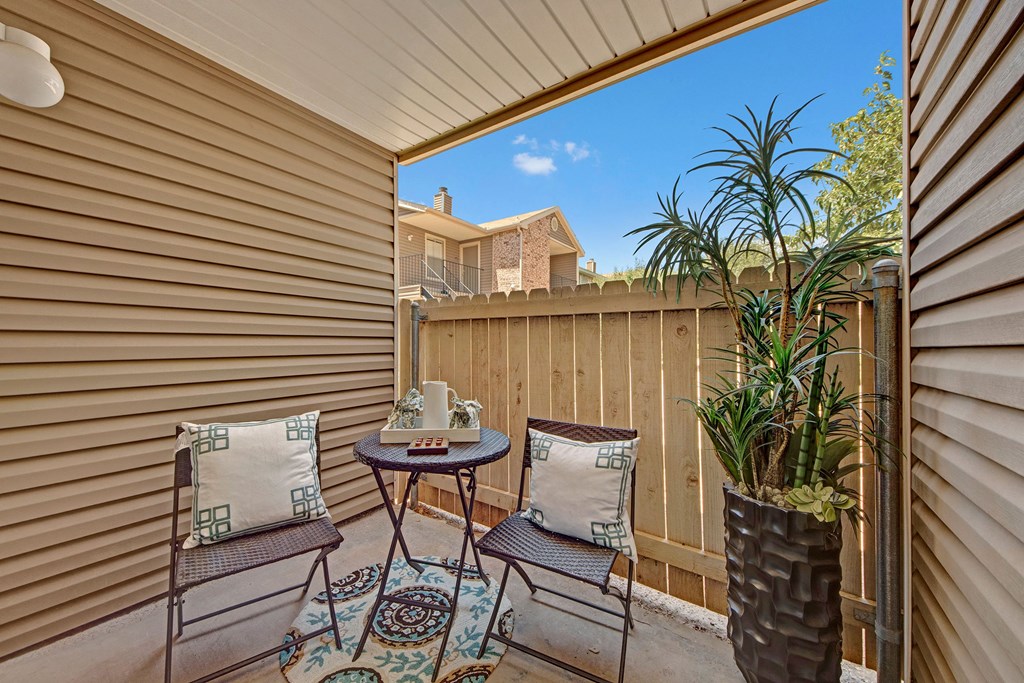 Cozy patio here at Northridge Court Apartment Homes with two chairs, patterned cushions, and a round table. Potted plant adds greenery. Wooden fence, house, and blue sky in the background create a relaxing vibe.