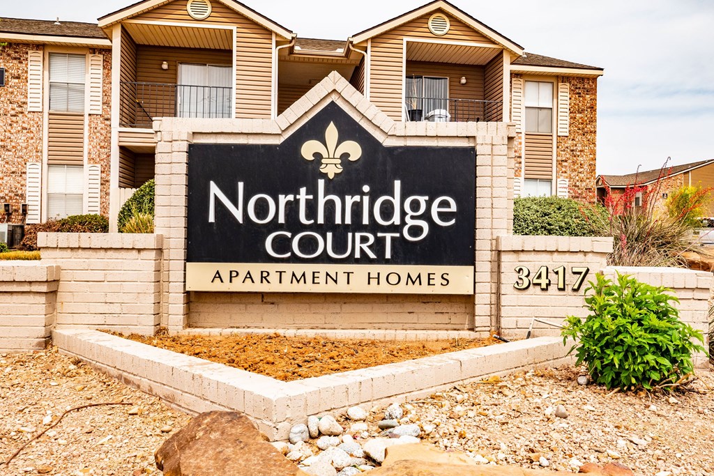 Sign reads "Northridge Court Apartment Homes" in front of brick buildings. Landscaping features rocks and shrubs. Bright, welcoming atmosphere.