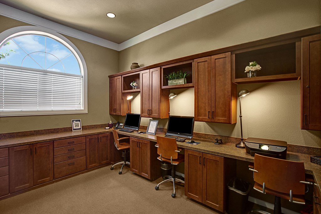 Warm-toned office space here at The Palms at Briarwood Apartment Homes with wooden cabinets, three workstations each with a computer, and a large arched window letting in soft natural light.
