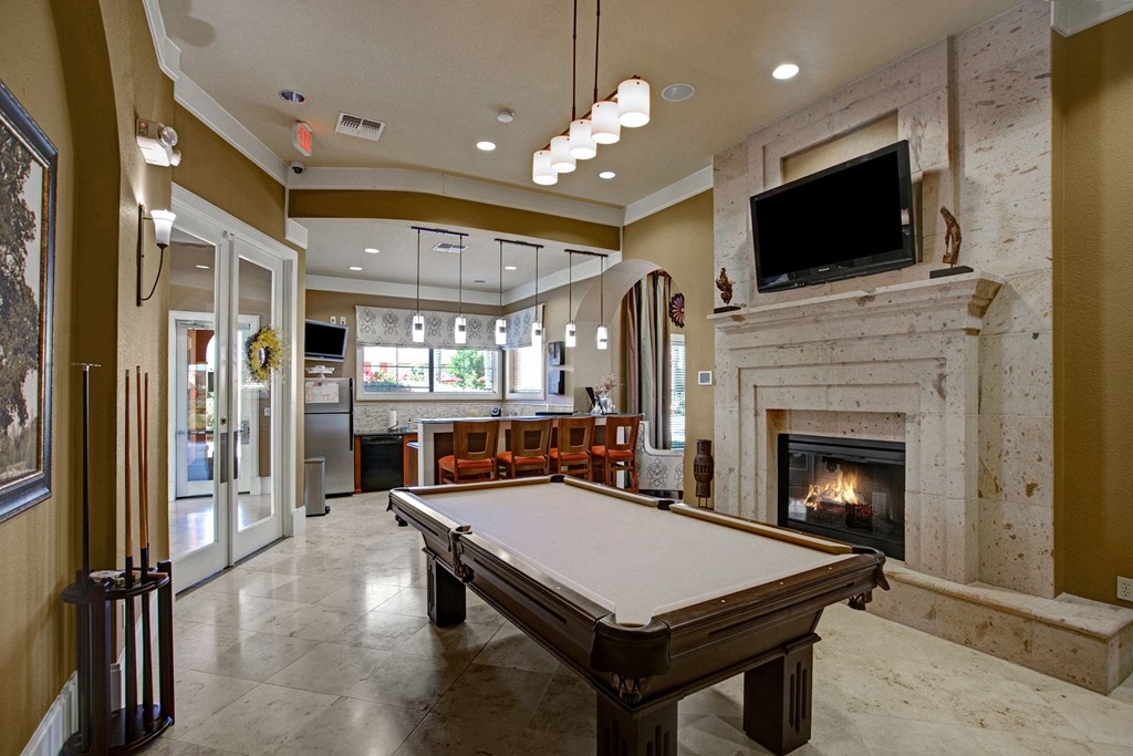 A cozy game room here at The Palms at Briarwood Apartment Homes with a pool table, beige walls, and marble floor. A lit fireplace and wall-mounted TV create a warm ambiance. A bar area with stools is visible in the background.