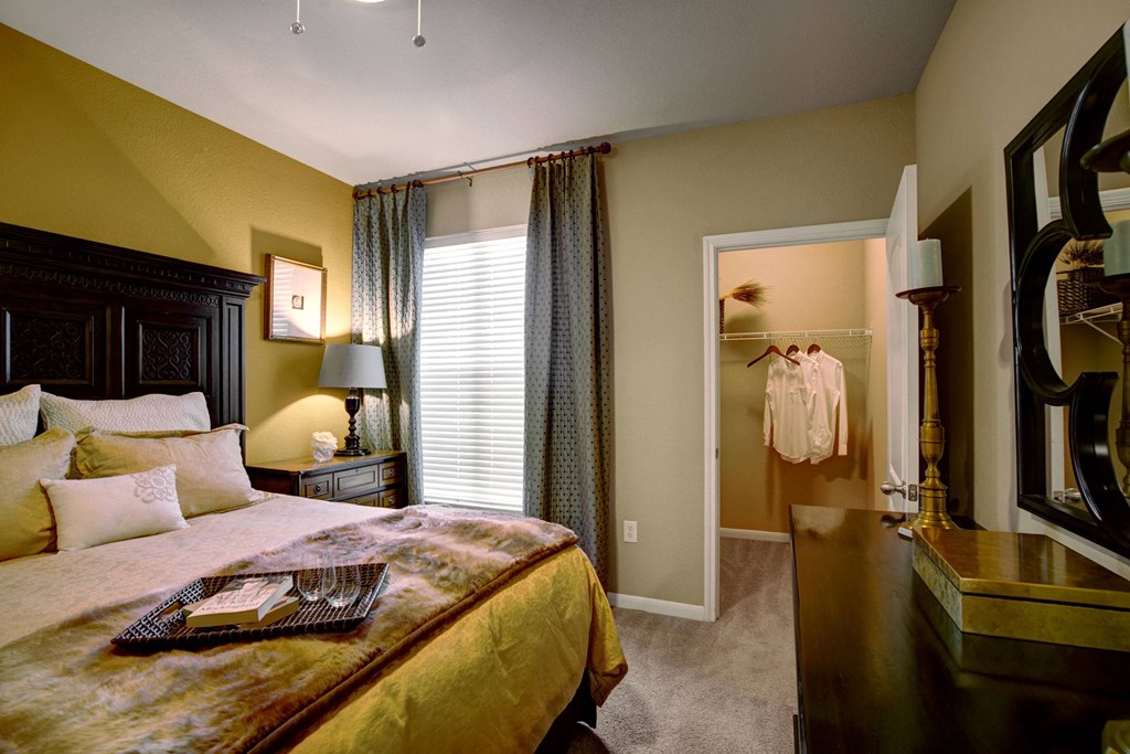 Cozy bedroom here at The Palms at Briarwood Apartment Homes with gold accents, dark wood furniture, and soft lighting. Bed has plush pillows and tray. Open walk-in closet shows a white shirt.