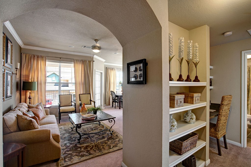 Cozy living room here at The Palms at Briarwood Apartment Homes with beige couch, patterned rug, and wooden coffee table. Shelving with decor and wicker chair in foreground. Warm lighting creates a welcoming atmosphere.