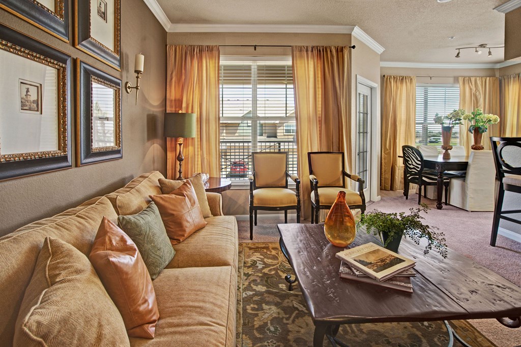 Cozy living room here at The Palms at Briarwood Apartment Homes with a beige sofa, orange cushions, wooden coffee table, and large windows with curtains. Warm, inviting atmosphere; dining area visible.