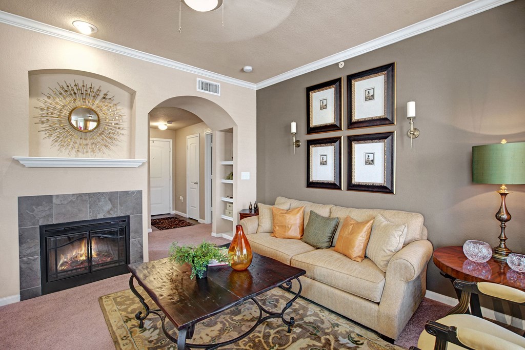 A cozy living room here at The Palms at Briarwood Apartment Homes with a beige sofa adorned with orange and green pillows. A modern fireplace and sunburst mirror add warmth. Art and a lamp enhance the elegant decor.