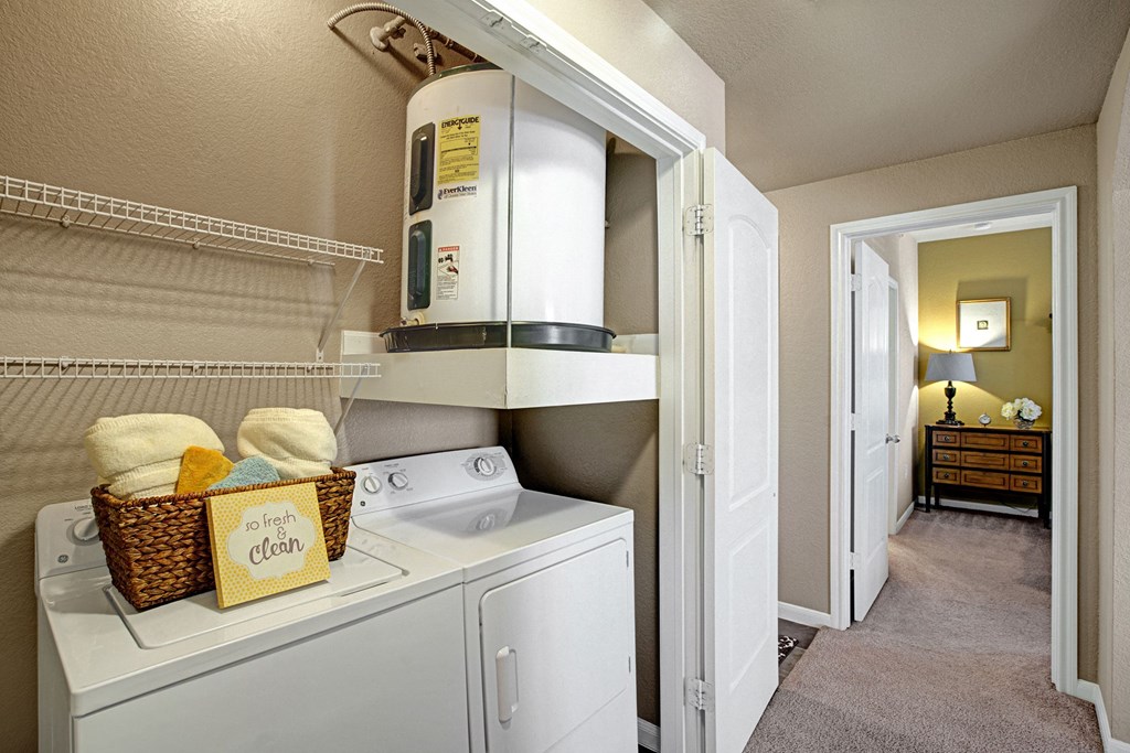 Laundry room here at The Palms at Briarwood Apartment Homes with washer, dryer, and water heater. A basket with folded towels sits on top, displaying a "So fresh & so clean" sign. Hallway view.