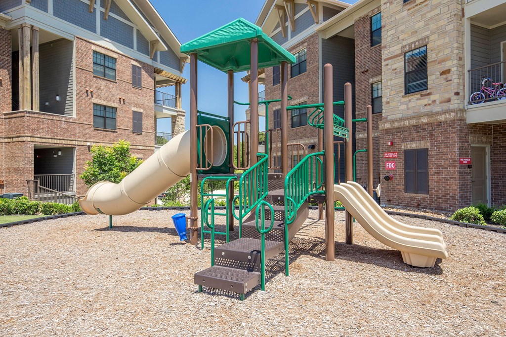 A vibrant playground here at Sorrel Fairview Apartment Homes with green and beige slides and climbing structures sits on wood chips. It's surrounded by modern brick apartment buildings under a clear blue sky.