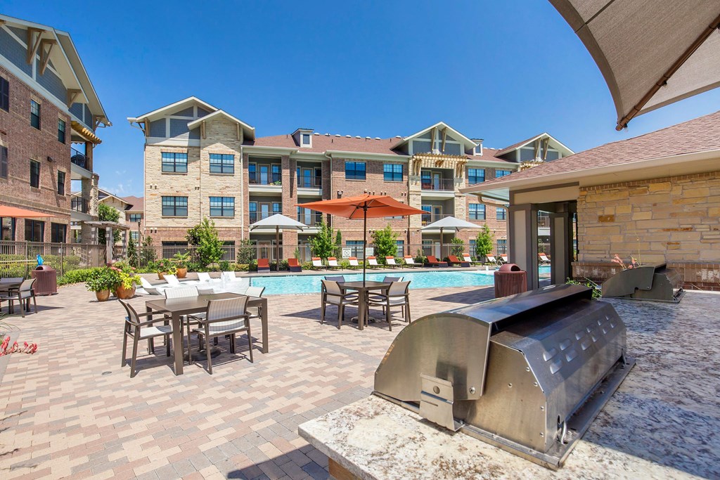 Modern courtyard here at Sorrel Fairview Apartment Homes with a pool, surrounded by lounge chairs and tables with umbrellas. A grill sits in the foreground under a clear blue sky.