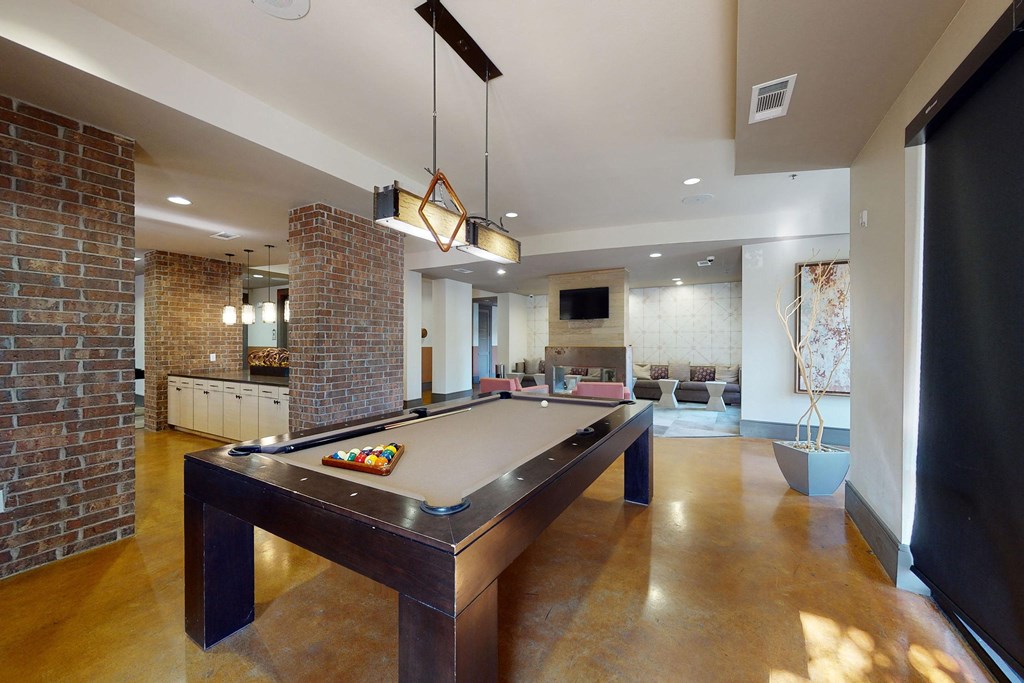 Luxurious recreation room here at Sorrel Fairview Apartment Homes with a brown pool table centered under a modern pendant light. Brick walls, cozy seating area with TV, and natural light add warmth.