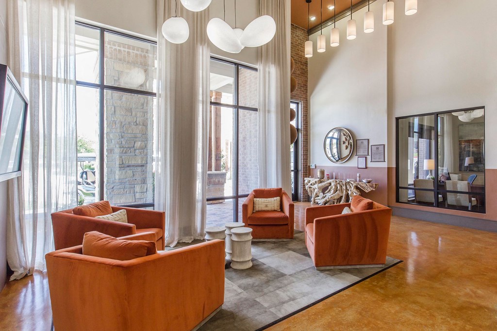 Modern living room here at Sorrel Fairview Apartment Homes with four orange armchairs on a gray rug, under oval pendant lights. Floor-to-ceiling windows and a neutral tone create a bright, cozy feel.