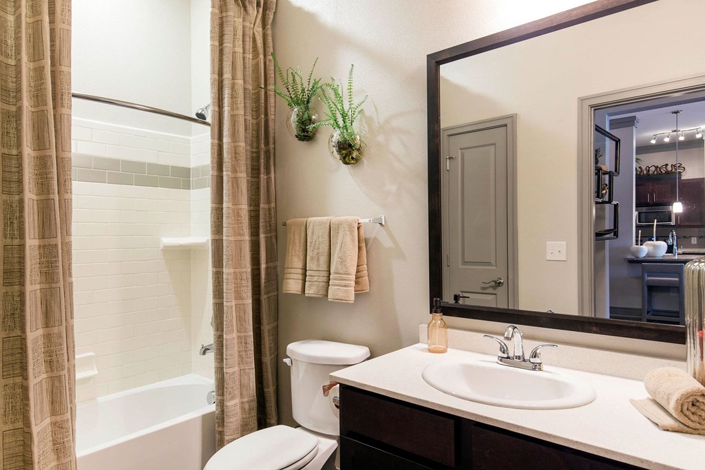 Modern bathroom here at Sorrel Fairview Apartment Homes with beige tiles and plaid shower curtain. A large mirror above a sink reflects a kitchen. Green plants and folded towels add decor.