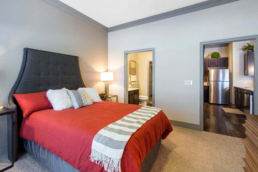 A cozy bedroom here at Sorrel Fairview Apartment Homes features a plush gray headboard, vibrant red bedding, and decorative pillows. Warm light from a bedside lamp enhances comfort. Doorways lead to a bathroom and kitchen with stainless steel appliances.