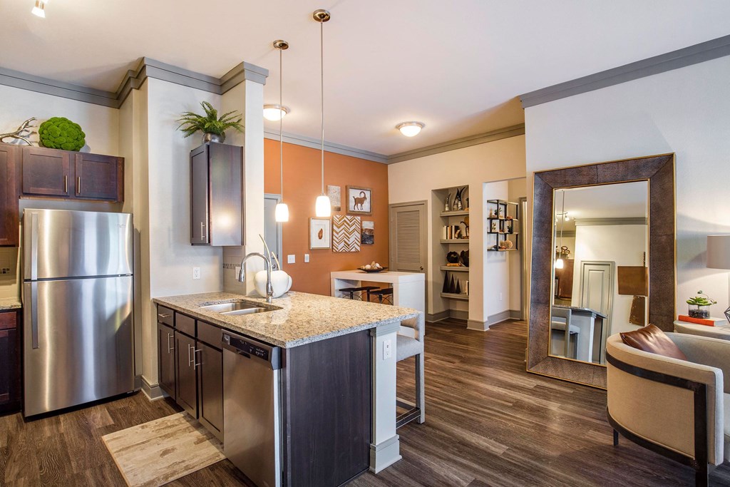 Modern kitchen here at Sorrel Fairview Apartment Homes with light wood floors, dark cabinets, stainless steel appliances, and pendant lighting. Cozy adjacent living area with a large mirror.