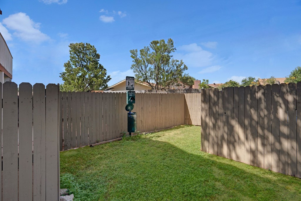 Fenced yard here at Summertree Place Apartment Homes with green grass and a pet waste station in the corner. Tall trees and a house are visible in the background under a clear blue sky.