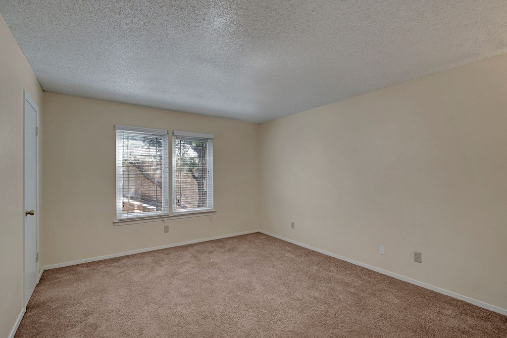 An empty room here at Summertree Place Apartment Homes with beige walls and carpet, a closed door on the left, and two large windows with blinds letting in natural light, creating a calm atmosphere.