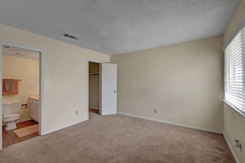 Empty carpeted room here at Summertree Place Apartment Homes with beige walls, an open closet, and a small bathroom visible. A window with blinds allows in soft light, creating a neutral, calm atmosphere.