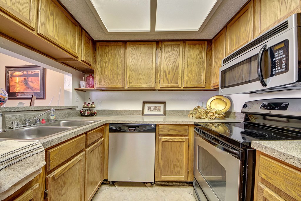 A well-lit kitchen here at Summertree Place Apartment Homes with wooden cabinets, stainless steel appliances, and a double sink. Decor includes a framed picture and a decorative plate. Cozy and organized.