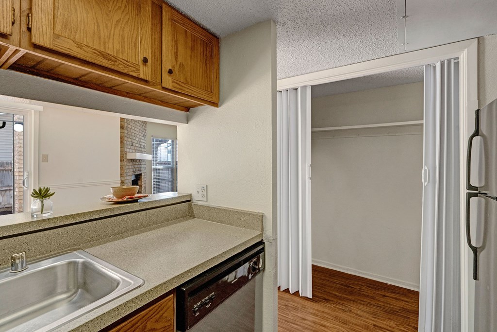 Small kitchen area here at Summertree Place Apartment Homes with brown cabinets, light countertops, and a stainless steel sink. There's an open closet with sliding doors. Tone is simple and neutral.