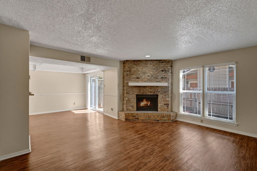 Spacious living room here at Summertree Place Apartment Homes with wooden floors, a brick fireplace, and large windows. Adjacent dining area features sliding glass doors, creating a cozy, inviting ambiance.