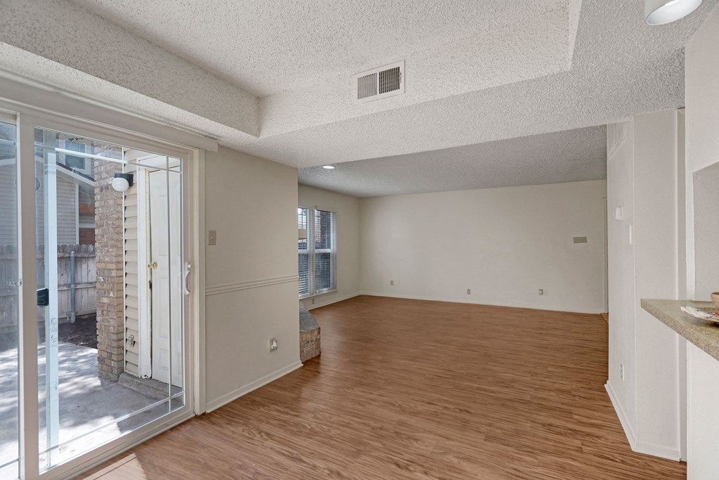 A spacious, empty living room here at Summertree Place Apartment Homes with wooden flooring and white walls, featuring a large window and a glass sliding door leading to a small patio. Bright and airy atmosphere.