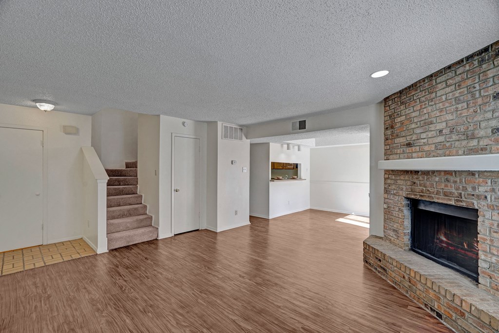 Empty living room here at Summertree Place Apartment Homes with wood floors, featuring a brick fireplace on the right. To the left are carpeted stairs, a small tiled area, and white walls. Bright and clean atmosphere.