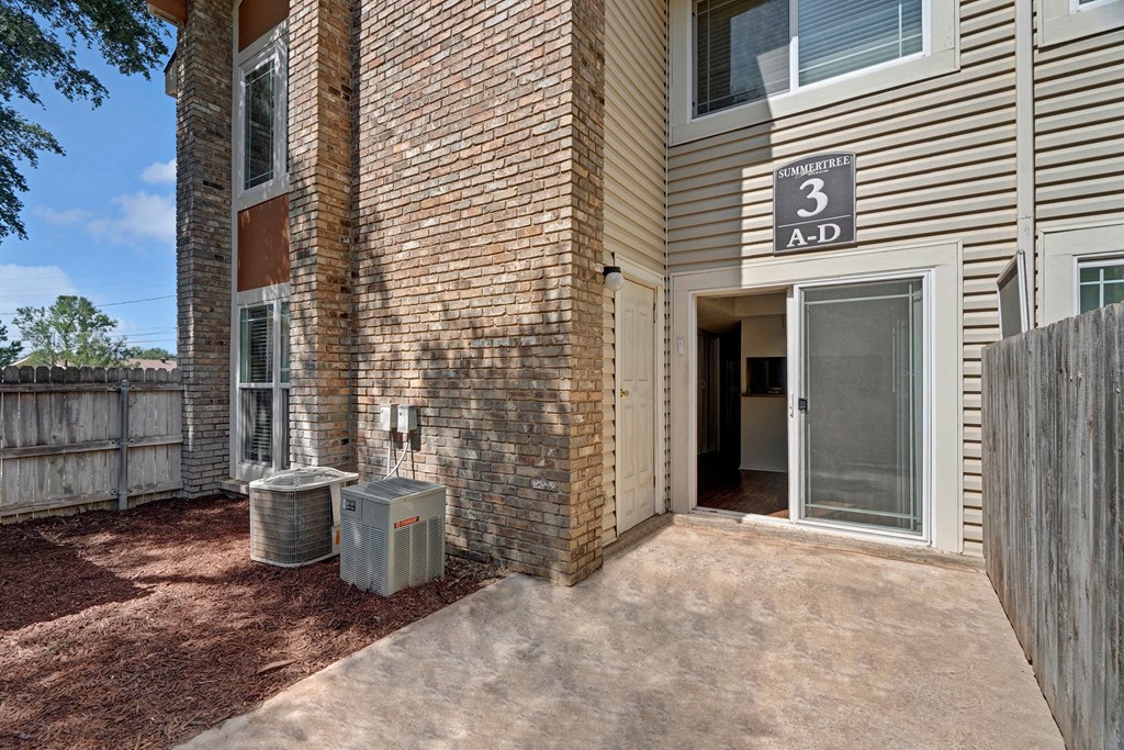 Exterior view of a residential building here at Summertree Place Apartment Homes with brick and siding. An open sliding door leads inside. Air conditioning units sit on red mulch, next to a wooden fence.