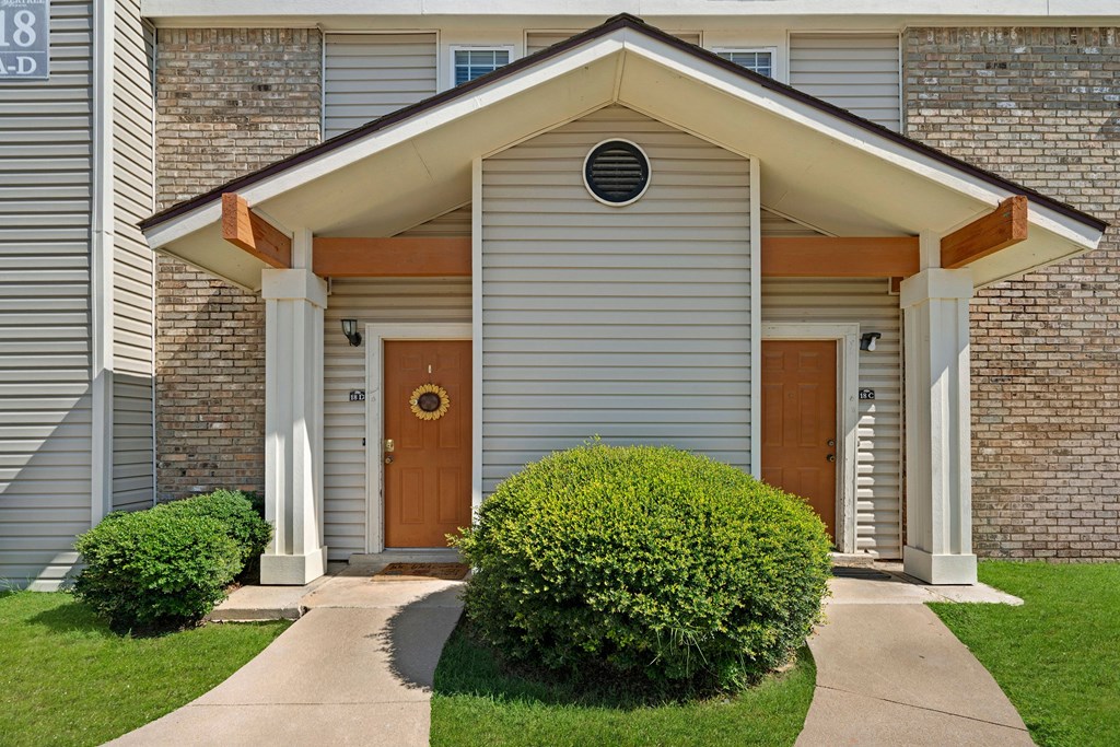 Front entrance of townhouses here at Summertree Place Apartment Homes featuring beige siding, brick accents, and two brown doors. A small gable extends overhead. Neatly trimmed bushes line the walkway.