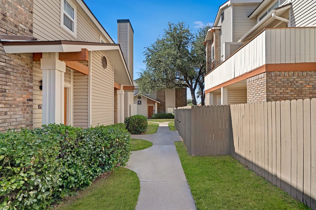 A narrow sidewalk runs between beige townhouses here at Summertree Place Apartment Homes with brick accents and wooden fences. Neat hedges line the path, and a large tree stands in the background. The scene is bright and serene under a clear blue sky.