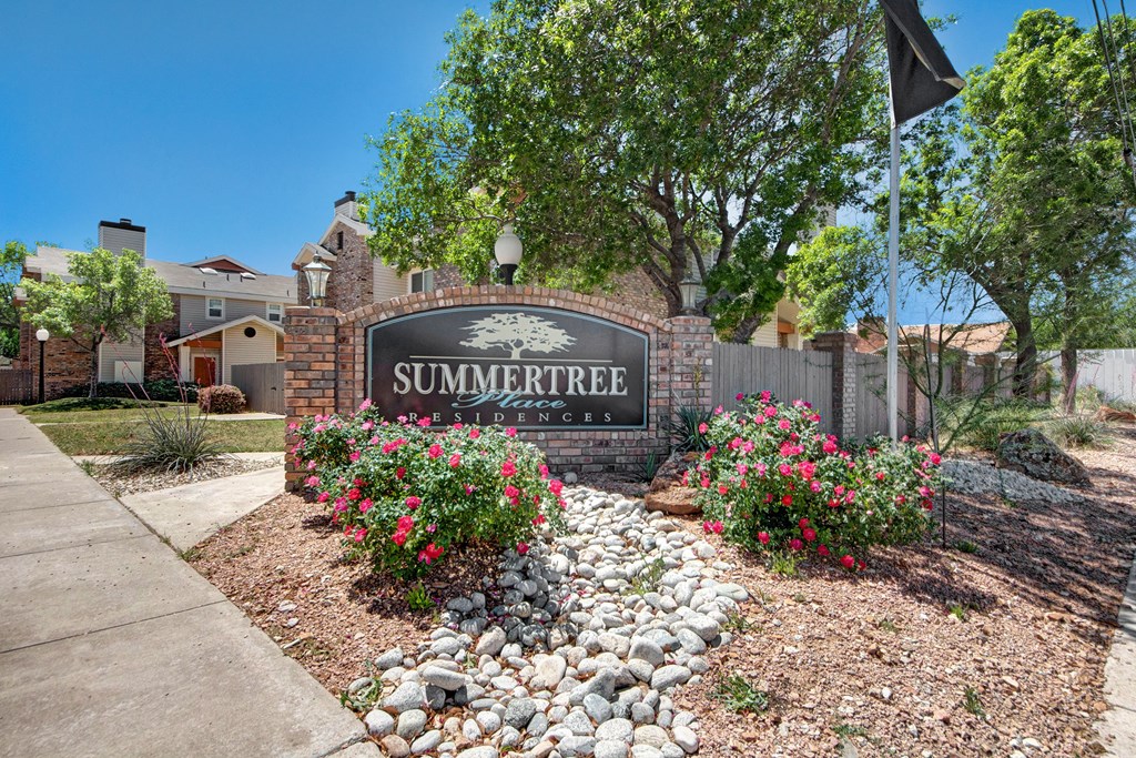 Entrance to Summertree Place Residences with a prominent sign surrounded by vibrant pink flowers and pebbles, set against a backdrop of trees and townhouses.