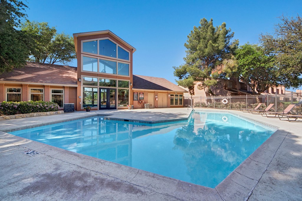 Outdoor pool here at The Bradford Apartment Homes beside a modern building with large windows under a clear blue sky, surrounded by trees and lounge chairs, conveying relaxation.