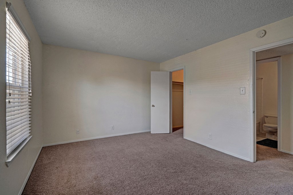 An empty bedroom here at The Bradford Apartment Homes with beige carpet and white walls. A window with blinds on the left, two open doors: one to a closet, the other to a bathroom. Bright and clean.