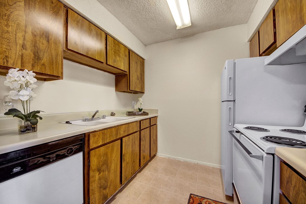 Compact kitchen here at The Bradford Apartment Homes with brown wood cabinets, white countertops, and appliances. Bright orchids in a clear vase add freshness. Neutral-tone floor and walls.